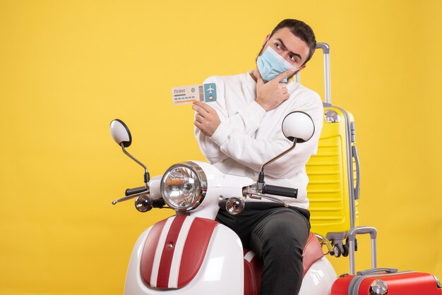 Top view of trip concept with thinking guy in medical mask sitting on motorcycle with yellow suitcase on it and holding ticket