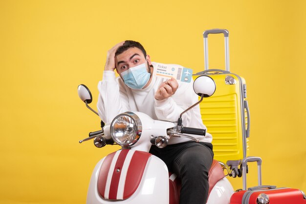 Top view of trip concept with young guy in medical mask sitting on motorcycle with yellow suitcase on it and holding ticket feeling confused