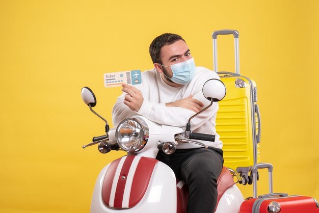 黄色いスーツケースを乗せたバイクに医療マスクを着た若い男が座り、驚きのチケットを持った旅行コンセプトのトップビュー