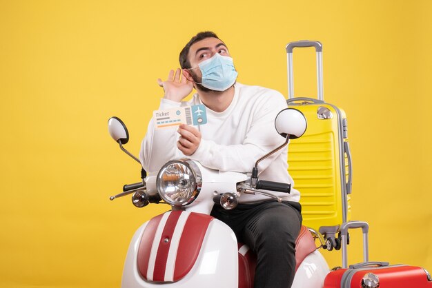 黄色いスーツケースを乗せたバイクに医療用マスクを着た若い男が座り、最後のうわさ話を聞いてチケットを持っている旅行コンセプトのトップビュー