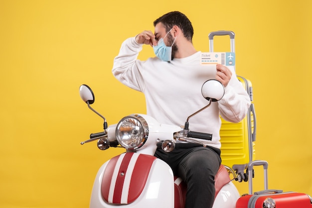 黄色いスーツケースを乗せたバイクに医療マスクを着た若い男が座り、悪臭のジェスチャーをするチケットを持った旅行コンセプトのトップビュー