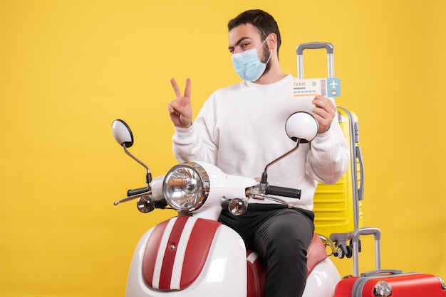 Vista dall'alto del concetto di viaggio con un giovane ragazzo in maschera medica seduto su una moto con valigia gialla su di esso e in possesso di biglietto facendo gesto di vittoria