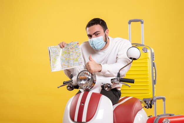 黄色いスーツケースを載せたバイクの近くに医療マスクを着た若い男が立ち、地図を持ってokのジェスチャーをする旅行のコンセプト
