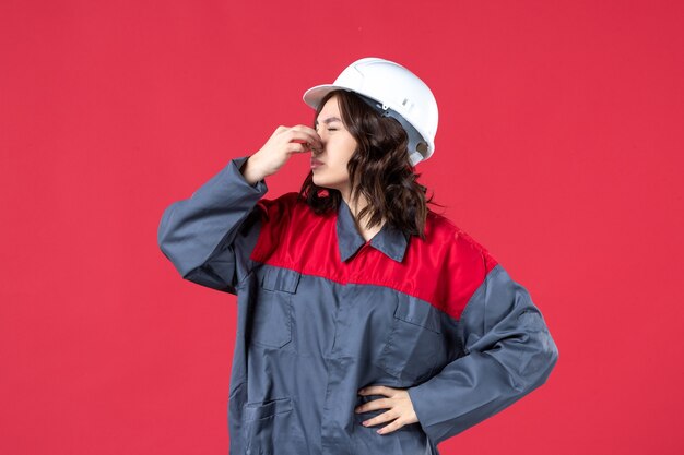 Top view of troubling female builder in uniform with hard hat and holding her nose on isolated red background
