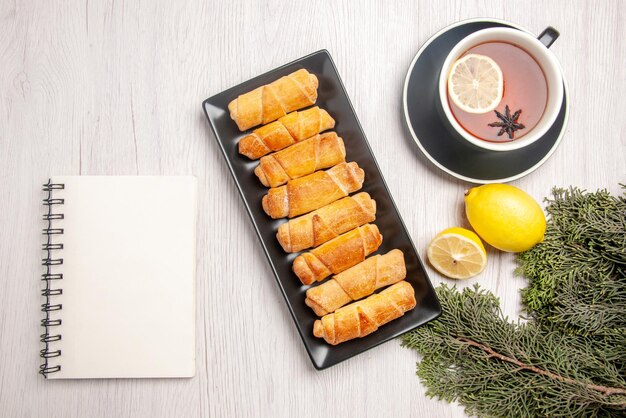 Top view tubular pastry tubular pastry on the black plate next to the white notebook cup of tea with lemon and spruce branches on the white table