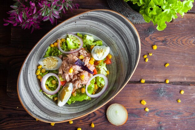 Салат из тунца с салатом, помидорами, яйцами, огурцом, луком и кукурузой на темной деревянной поверхности.