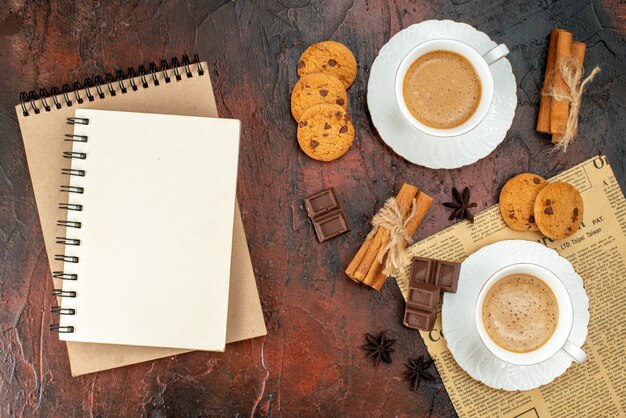 Top view of two cups of coffee cookies cinnamon limes chocolate bars on an old newspaper and notebooks on dark background