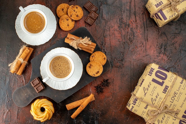 Top view of two cups of coffee cookies cinnamon limes chocolate bars on wooden cutting board and gift boxes on dark background