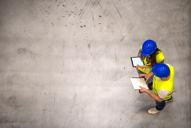 Top view of two industrial workers wearing hardhats and reflective jackets holding tablet and checklist on gray concrete floor