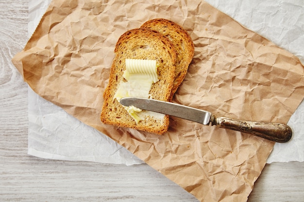 ヴィンテージナイフを載せた朝食用のバター付きトーストとしてのライ麦ドライパンの2つのスライスの上面図。クラフト紙のすべて。