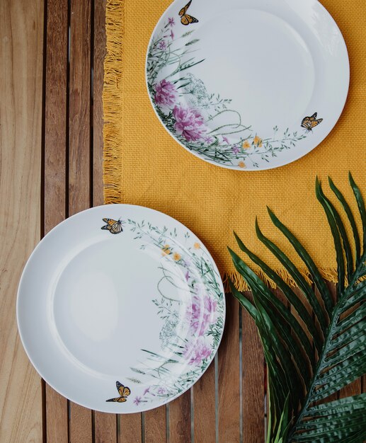 Top view of two table empty plates with flower pattern on a yellow napkin on wooden wall