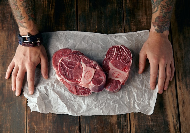 Top view of two tattooed hands and three steaks on white craft paper on old wooden table