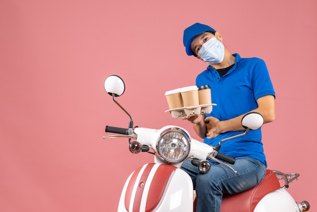 Top view of uncertain courier man in mask wearing hat sitting on scooter showing orders on pastel peach