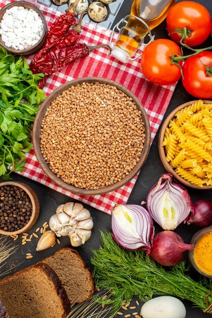 Top view of uncooked buckwheat in a brown bowl on towel fresh vegetables fallen oil bottle salt green bundle dried pepper