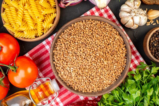 Top view of uncooked buckwheat in a brown bowl on towel pasta in pot fresh vegetables fallen oil bottle salt green bundle bread slices