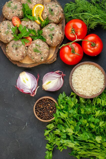 Top view of uncooked meatballs on a wooden cutting board and fresh vegetables pepper salt on dark wave background