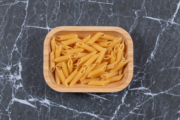 Top view of uncooked pasta penne in wooden bowl.