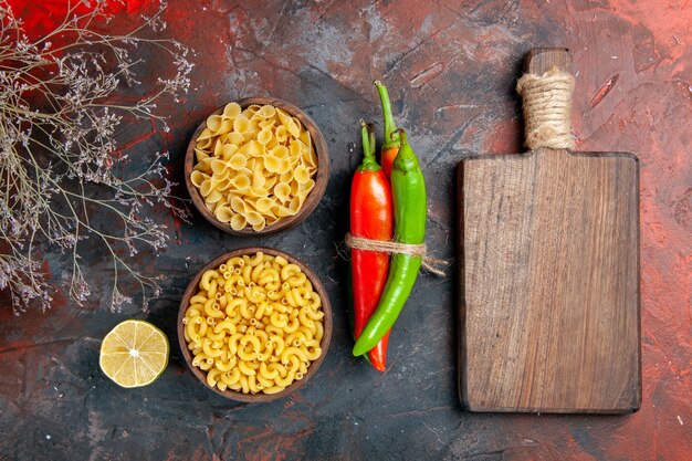 Top view of uncooked pastas cayenne peppers in different colors and sizes tied in one another with rope and wooden cutting board on mixed color background