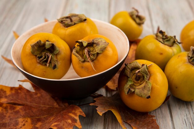 Top view of unripe persimmon fruits on a bowl with leaves on a grey wooden table