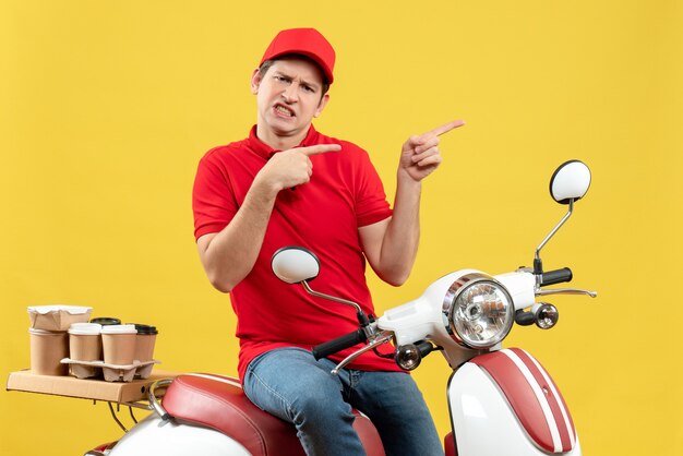 Top view of unsatisfied young guy wearing red blouse and hat delivering orders pointing something on yellow wall