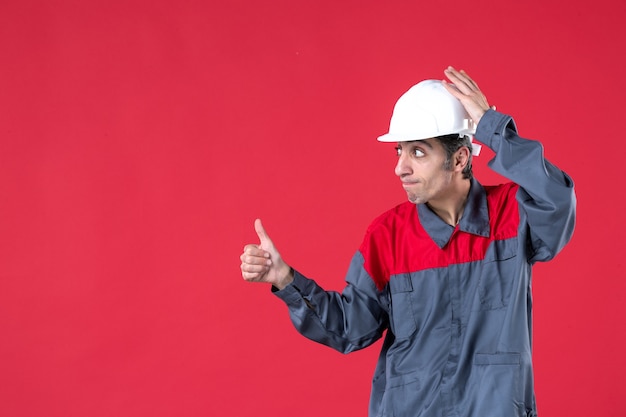 Free photo top view of unsure young worker in uniform with hard hat pointing up and making ok gesture on isolated red wall