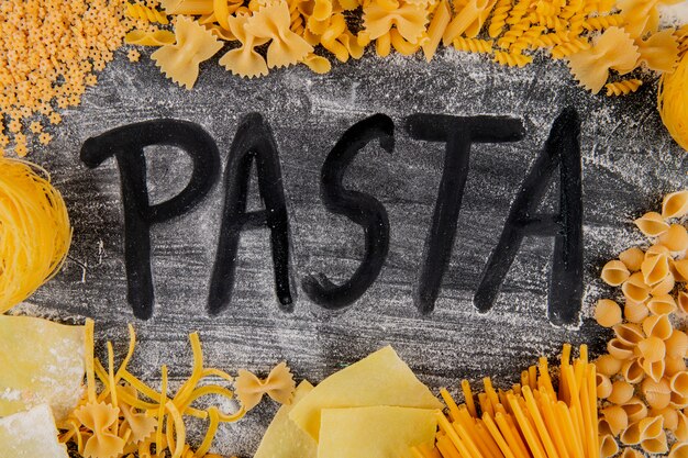 Top view of variety of types and shapes of italian pasta and word pasta from flour on dark background