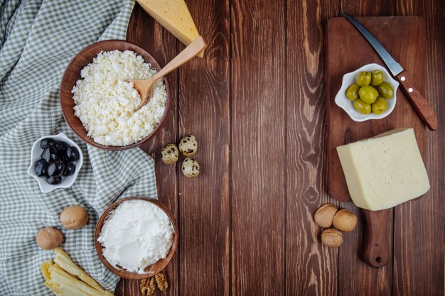 Top view of various cheese and cottage cheese in a bowl with walnuts, quail eggs and pickled olives on wooden cutting board with a knife on rustic table