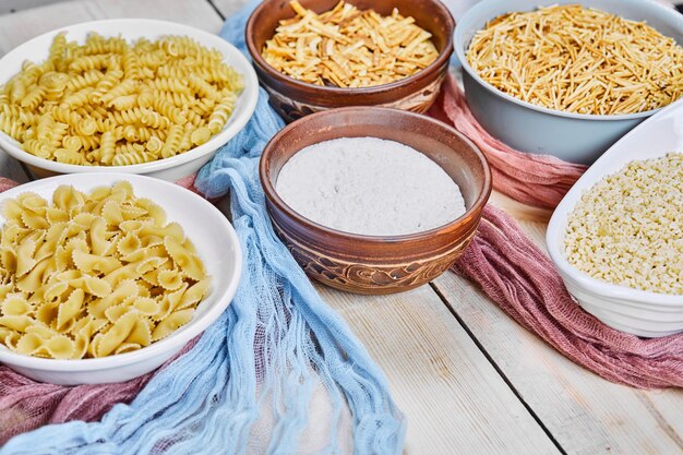 Top view of various raw pastas and bowl of flour on wooden table with blue and pink tablecloth.