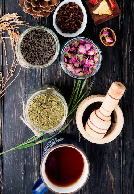 Top view of various spices and herbs dry black tea leaves, peppermint, rose buds, clove spice and black peppercorns in glass jars on black wood