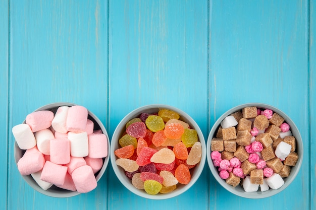 Top view of various sweets colorful marmalade candies marshmallow and brown sugar cubes in bowls on blue