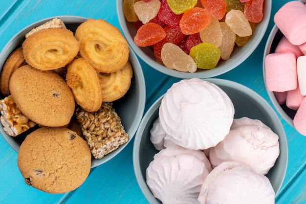 Top view of various sweets cookies colorful marmalade candies and white zephyr marshmallows in bowls on blue