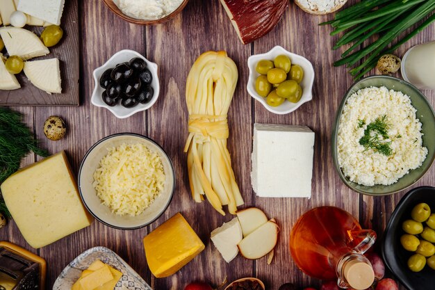 Top view of various types of cheese with green onio honey in a glass bottle and pickled olives on rustic wood