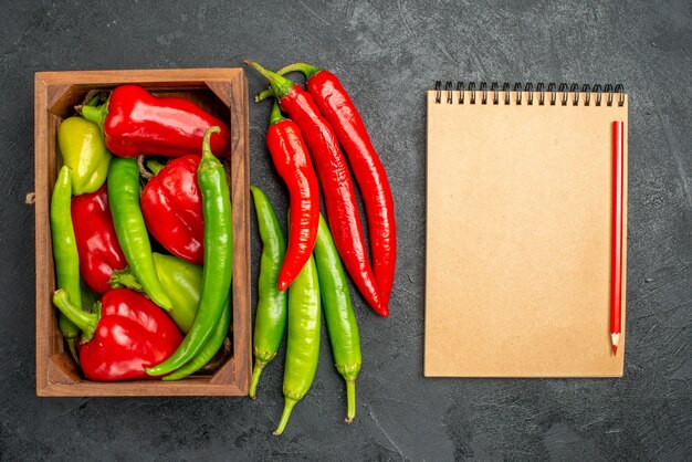 Top view various types of peppers inside the box near to notebook and red pen on black surface with free space