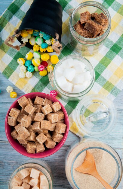 Top view of various types of sugar in glass jars and colorful sugar candies scattered from a bucket on plaid table napkin on rustic background