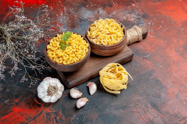 Top view of various types of uncooked pastas on wooden cutting board and garlic on mixed color background