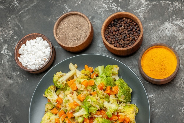 Top view of vegetable meal with brocoli and carrots on a black plate and spices on gray background