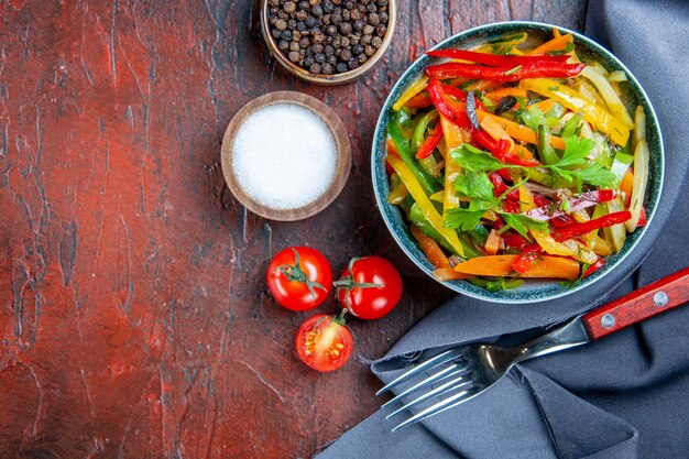 Top view vegetable salad in bowl ultramarine blue shawl cherry tomatoes spices fork on dark red table free space