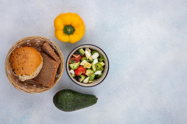 Top view of vegetable salad including cucumber tomato pepper in a bowl with a basket of breads yellow bell pepper and avocado on white surface