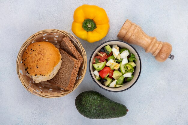 Top view of vegetable salad including tomato cucumber pepper in a bowl with a bucket of bread yellow bell pepper and avocado on white surface