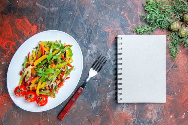 Top view vegetable salad on oval plate fork notebook pine branches on dark red table