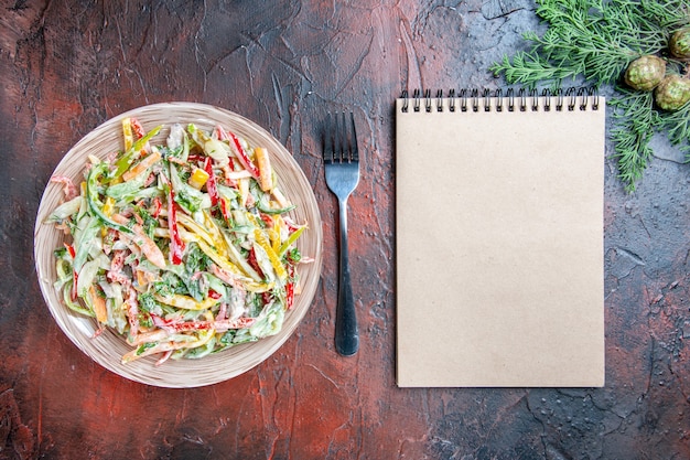 Free photo top view vegetable salad on plate fork notepad on dark red table