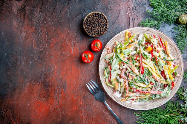 Top view vegetable salad on plate fork tomatoes pine branches on dark red table free space