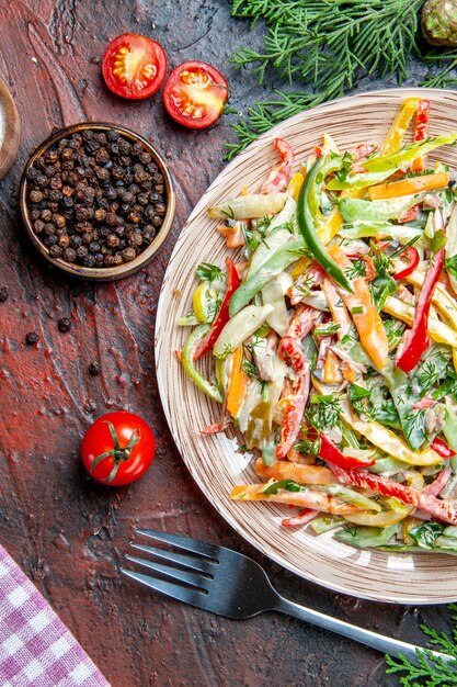 Top view vegetable salad on plate tablecloth fork black pepper tomatoes on dark red table