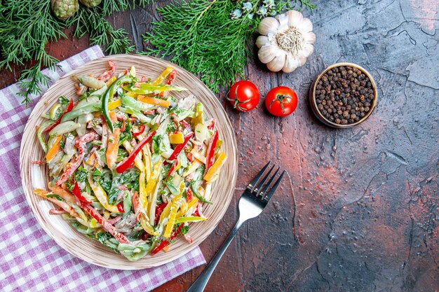 Top view vegetable salad on plate on tablecloth fork black pepper tomatoes garlic on dark red table