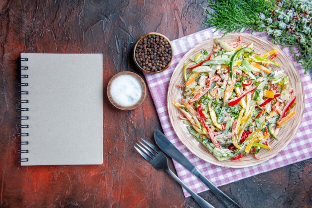 テーブルクロスのフォークとナイフの塩と黒コショウのプレート上のトップビュー野菜サラダ濃い赤のテーブルの上のノート