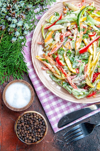 Вид сверху овощной салат на тарелке на скатерти, вилка и нож, соль и черный перец, сосновые ветки на темно-красном столе