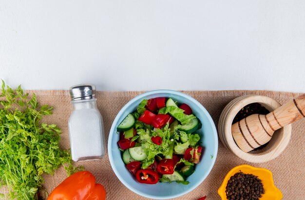 Top view of vegetable salad with bunch of coriander pepper salt black pepper seeds on sackcloth and white surface with copy space