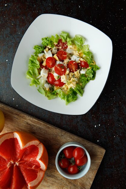 Top view of vegetable salad with tomato egg lettuce in plate with cut grapefruit and bowl of tomatoes on black background