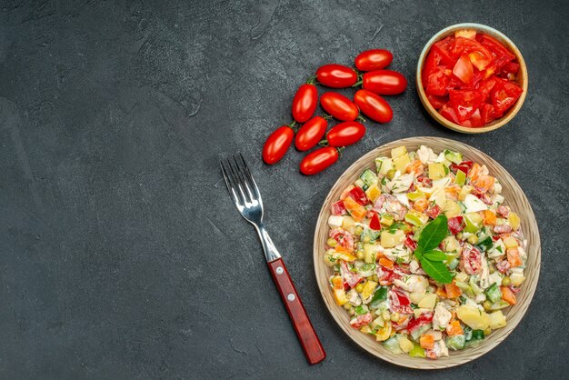 Top view of vegetable salad with tomatoes and fork on dark grey background