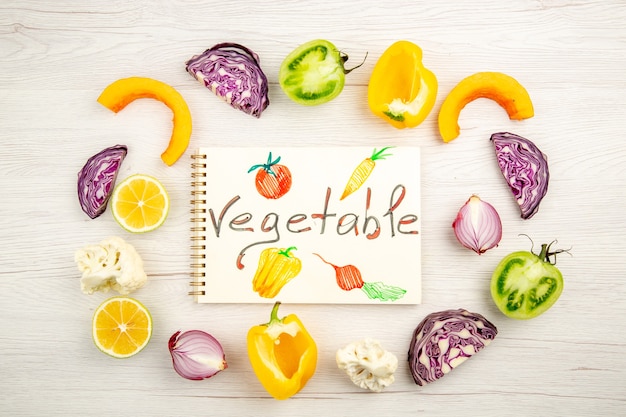 Top view vegetable written on notepad cut vegetables red cabbage green tomato pumpkin red onion yellow bell pepper caulifower lemon on white table